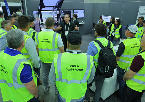 A group of people wearing bright yellow safety vests labeled 'Field Classroom' gathered around a demonstration area with large screens and equipment in an indoor setting.